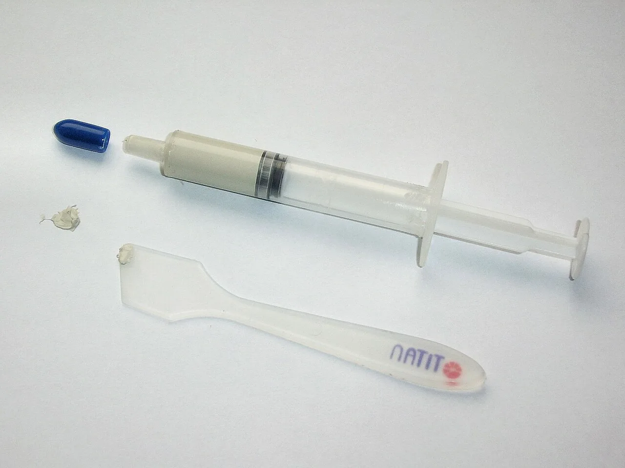 Thermal paste syringe with its plastic spreading applicator and a small blob of grey compound on a white surface